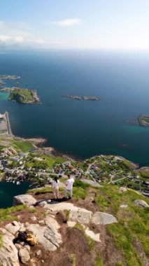 İki yürüyüşçünün bir dağ zirvesinden panoramik manzaranın keyfini çıkarırken Norveç 'in huzurlu güzelliği. Manzara, berrak bir gökyüzü altında yemyeşil ve ışıl ışıl sulara sahip, Reinebringen Lofoten Norveç yürüyüşü