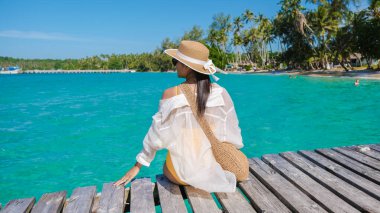 Koh Kood, Tayland 'ın canlı mavi sularında huzurlu bir an. Bir kadın, bereketli palmiyeler ve açık bir gökyüzü ile çevrili, ahşap bir iskelede oturan tropikal güneşin tadını çıkarır..