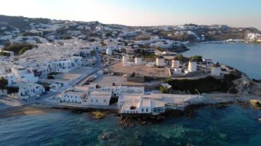 Mikonos Yunanistan 'ın sakin kıyı şeridine bakan ikonik yel değirmenleriyle güzelliğini keşfedin. Güneş, beyaz mimariyi ve parlak mavi denizi aydınlatan pitoresk bir manzaranın üzerinde batar..