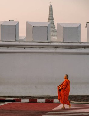 10 Ocak 2025 'te Bangkok Tayland' da parlak turuncu giyinmiş bir keşiş yaya geçidinde yürürken alacakaranlığın yumuşak renkleriyle aydınlatılan görkemli tapınak arka planda beliriyor..