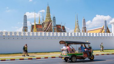 10 Ocak 2025 'te Bangkok Tayland' da, parlak mavi bir gökyüzünün altında, muhteşem Wat Phra Kaew tapınağının yanından renkli tuk-tuklar süzülüyor. Turistler hareketli sokaklarda dolaşıyorlar.