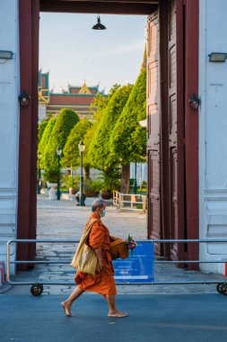 Bangkok Thailand 10 January 2025, A monk in vibrant orange robes walks gracefully through traditional gates, carrying offerings. captures the essence of a peaceful morning at the Grand Palace. clipart