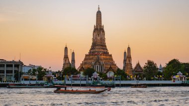 Güneş batarken, Wat Arun Bangkok 'taki Chao Phraya Nehri üzerinde görkemli kuleler inşa eder. Tekneler tembelce geçip giderken, tapınağın karmaşık mimarisi alacakaranlığın sıcak tonlarını yansıtıyor..