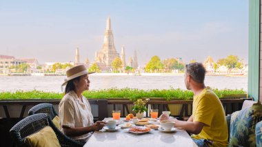 Bangkok 'ta nehir kıyısında sakin bir kahvaltı. Bir çift lezzetli yemeğin tadını çıkarırken ikonik Wat Arun tapınağı ve yemyeşil çevresinin nefes kesici manzarasını izliyorlar..