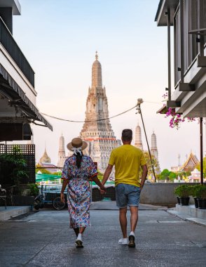 A couple enjoys a leisurely walk hand in hand down a charming street in Bangkok, approaching the stunning Wat Arun. The evening light bathes the scene in warmth and romance. clipart