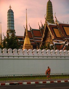 10 Ocak 2025 'te Bangkok Tayland' da, turuncu cüppeli bir keşiş gün batımında karmaşık mimari üzerine sıcak bir parıltı saçan Grand Royal Palace, MajesticWat Phra Kaew Tapınağı yakınlarında sessizce yansıyor.