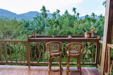 Bir çift tahta sandalye, yemyeşil bitkilerle süslenmiş büyüleyici bir balkonda rahatlamaya davetiye çıkarır. Aşağıda, canlı yeşillik uzak dağlara doğru yayılıyor, Tayland 'ın doğal güzelliklerini yansıtıyor..