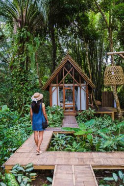 Bir kadın, canlı yaprakların arasına yerleştirilmiş büyüleyici bir kulübeye doğru çekilen ahşap bir patikada geziniyor. Huzurlu ortam, yağmur ormanlarında doğayla olan rahatlığı ve bağlantıyı davet eder.