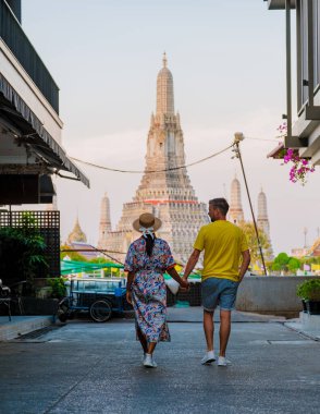 İkonik Wat Arun arka planda yükselirken bir çift el ele büyüleyici bir sokakta yürüyor. Gün batımının ılık ışığı Bangkoks sokaklarının canlı atmosferini güçlendirir..