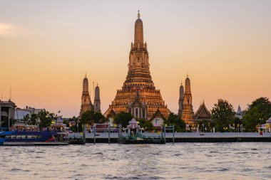 Wat Arun, gün batımında Bangkok 'ta ihtişamlı bir şekilde aydınlanarak Chao Phraya Nehri' ni yansıtıyor. Gemiler yavaşça süzülüyor gökyüzü ılık renklerle dönüşürken.