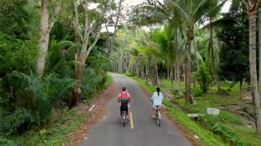 Chumphon Tayland 'da canlı tropikal yapraklarla çevrili sakin bir patika boyunca bisiklet sürerken, arkadaşlar parlak bir gökyüzü altında bölgeyi keşfediyorlar. Bir çift bisikletli kadın ve erkek.