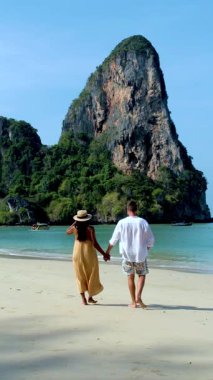 Bir çift, arkalarında görkemli bir kireçtaşı uçurumu yükselirken Tayland, Railay Beach Krabi 'nin kumlu sahillerinde romantik bir yürüyüş yapıyor. Berrak turkuaz sular ve yemyeşil bolluk.