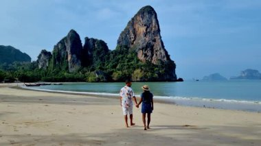 Railay plajı boyunca el ele yürürken Krabi Tayland 'da nefes kesici kaya oluşumları ve berrak mavi sular görülür. Çiftler bu tropik cennetin huzurlu atmosferinin tadını çıkarıyorlar..