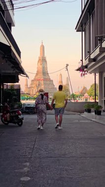 Gün batımında Bangkok 'un canlı sokaklarında el ele yürüyoruz. Arka plandaki görkemli Wat Arun tapınağı bu büyüleyici şehirde büyülü bir atmosfer yaratıyor..