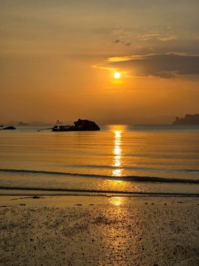 Güneş Krabi Tayland 'ın arkasında batarken, gökyüzü durgun sulara yansıyan canlı turuncu renklerle dönüşür. Kıyı sönen ışıkta parıldarken bir tekne huzur içinde sürüklenir..