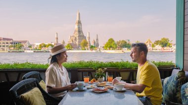 İki arkadaş Bangkok 'ta nehir kenarındaki terasta keyifli bir kahvaltıyı paylaşıyor. İkonik Wat Arun tapınağı açık bir gökyüzünün altında ihtişamlı bir şekilde yükselir ve güzel bir sabah anı yaratır..