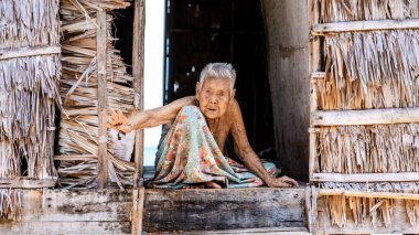 Phangnga, Tayland, 21 Ocak 2025, Surin Adası 'ndaki geleneksel evinin girişinde rahat bir şekilde oturan yaşlı bir kadın. Etrafı bereketli doğayla çevrili, rahatlatıcı kıyı melteminin tadını çıkarıyor.