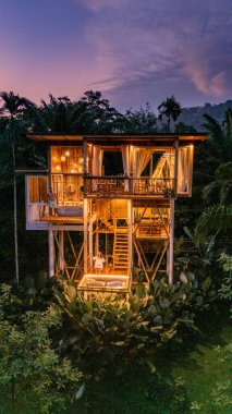 Tayland 'da canlı yeşilliklerin arasına yerleştirilmiş güzel tasarlanmış bir ağaç ev huzurlu bir kaçış sunuyor. Alacakaranlık yaklaşırken, sıcak ışıklar yapıyı aydınlatıyor ve büyüleyici cazibesini arttırıyor..
