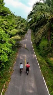İki bisikletçi, Chumphon Thailand 'da dolambaçlı bir yol boyunca, canlı yeşillikler ve uzun palmiye ağaçlarıyla çevrili, huzurlu bir yolculuğun tadını çıkarıyorlar..