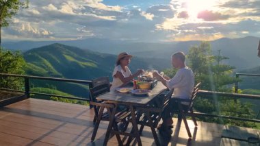 Çift, Tayland, Chiang Mai 'nin büyüleyici pirinç tarlalarına bakarken keyifli bir yemek yiyor. Günbatımı gökyüzünü canlı renklerle boyar, romantik bir atmosfer yaratır..