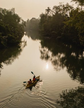 İki maceracı Chumphon 'un sakin sularında, Tayland' da palmiye ağaçlarıyla çevrili bir kanoyla, yemyeşil çimlerle çevrili bir kanoyla zarifçe kürek çekerler..