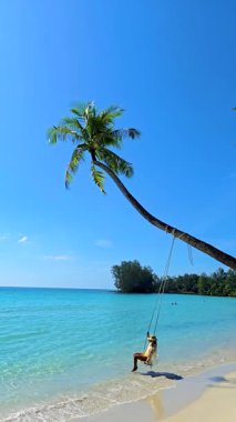 Yumuşak dalgalar kıyıya vururken Koh Kood Adası 'nın huzurlu güzelliğini hissedin. Plajda sallanan bir kadın bir palmiye ağacının altında, berrak mavi gökyüzü ve okyanusun nefes kesici manzarasının tadını çıkarıyor..