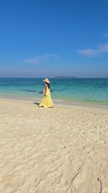 Bir kadın kıyı boyunca zarifçe yürürken parlak güneş ışığı yumuşak kumu aydınlatır. Kristal berrak sular nazikçe ayaklarına dolanıyor, Koh Munnork Adası Tayland 'da sakin bir atmosfer yaratıyor..