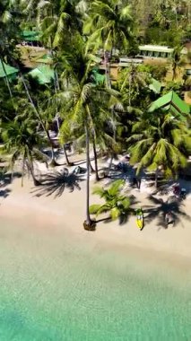 Koh Kood Tayland 'daki sakin plajda yumuşak kumun üzerine düşen yemyeşil palmiye ağaçları yer alıyor. Ziyaretçiler sakin atmosferin ve çarpıcı mavi suların tadını çıkarıyorlar.. 