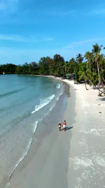 Tayland 'daki Koh Kood Adası' nın el değmemiş kumlu sahillerinde iki kişi el ele yürüyor. Bereketli palmiye ağaçları rüzgarda hafifçe sallanır, sakin bir atmosfer yaratır..