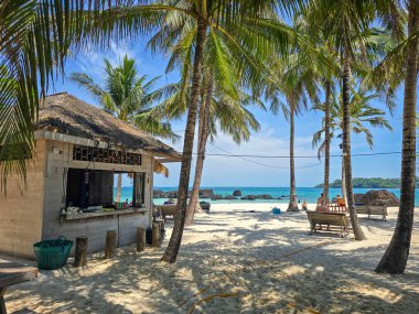 Koh Kham Adası 'ndaki çarpıcı sahil manzarası toz beyazı kum, gür palmiye ağaçları ve büyüleyici bir sahil kulübesi sergiliyor. Ziyaretçiler güneşin altında dinleniyor, Tayland 'ın sakin tropik cennetinin tadını çıkarıyorlar..