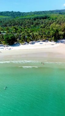Tayland 'ın Koh Kood Adası' ndaki el değmemiş kumlu bir plajda turkuaz su turkuazlarını sakinleştirir. Bereketli yeşil palmiye ağaçları, ziyaretçilerin güneşin altındaki dingin atmosferin tadını çıkardıkları sakin bölgenin etrafını sarar..