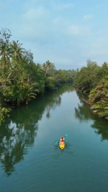 Tayland 'ın sakin bir nehrinde canlı palmiyeler ve yemyeşil bitkilerle çevrili bir kürekçi sorunsuzca süzülür. Su aktivitelerinin tadını çıkarırken doğanın güzelliğini hissedin..
