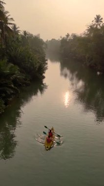 Sabahın erken saatlerinde Chumphon Tayland 'da sakin sularda kürek çeken bir çift çarpıcı bir nehir manzarasını gözler önüne seriyor. Siluetli kano, yemyeşil bitki örtüsünün yanında süzülür şafağın yumuşak ışığını yansıtır..