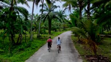 İki bisikletçi, Tayland 'ın verimli kırsallarında, canlı palmiyelerle çevrili sakin bir yolda ilerliyorlar. Barışçıl atmosfer macera ve doğa keşfine davet ediyor güzellik..