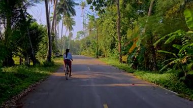 Chumphon Thailand 'ın sakin güzelliğini keşfedin. Bir kadın bisikletçi virajlı bir yolda giderken çevreleyen yeşil ve uzun palmiye ağaçları kırsal sükunetin özünü yakalar.