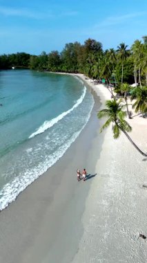 Tayland 'daki Koh Kood Adası' nın güzelliğini tecrübe edin. İki insan el değmemiş kumsalda, yemyeşil ve parlak mavi bir gökyüzü altında hafif dalgalarla dolu huzurlu bir gezinti yaparken..