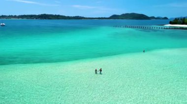 Tayland 'daki Koh Kham Adası' nın nefes kesici güzelliğini deneyimleyin. Yumuşak beyaz kumların berrak turkuaz sularla buluştuğu yer. Ziyaretçiler kumsalda gezinir, cennette huzurlu anların tadını çıkarırlar..