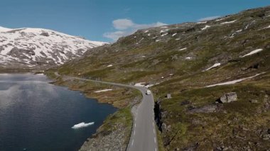 Norveç 'in nefes kesici manzaralarını sakin sularla çevrili dolambaçlı bir yol ve berrak mavi gökyüzünün altındaki görkemli kar örtülü zirvelerle keşfedin. Geiranger Norveç yakınlarındaki Langvatnet yolunda bir kamp aracı.