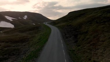 Kuzey Burnu 'nun Norveç görkemli dağları içinde, yemyeşil ve kar kalıntılarıyla çevrili sakin, dolambaçlı bir yol keşfedin. Batan güneş, manzaranın üzerine sıcak bir ışık saçıyor..