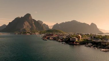 Lofoten Norveç 'te dramatik dağlarla çevrili bir sahil köyünün huzur veren güzelliğini görün. Altın gün batımı, sakin suların ve büyüleyici mimarinin üzerine ılık bir ışık saçıyor..