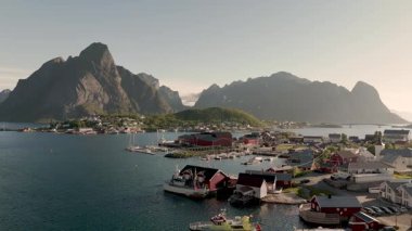 Norveç 'in nefes kesen kıyı köyünde etkileyici dağların arka planına kurulmuş geleneksel kırmızı balıkçı kulübeleri ve tekneler sergileniyor. Reine Lofoten