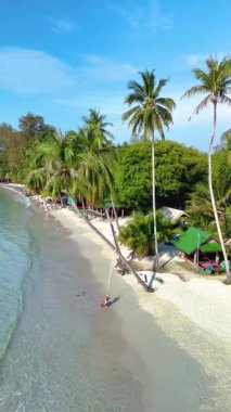 Koh Kood Adası 'ndaki sakin plajda yumuşak kumlu kıyıya eğilmiş palmiye ağaçları var. Kristal berrak sular mavi gökyüzünü yansıtıyor ziyaretçiler Tayland 'ın sakin atmosferinin tadını çıkarırken