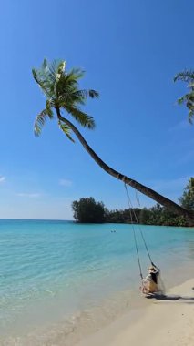 Koh Kood Tayland 'da sakin bir sahil sahili, yumuşak beyaz kumun üzerinde yavaşça sallanan uzun bir palmiye ağacına asılı bir salıncağa sahiptir. Turkuaz su, parlak güneşin altında parlıyor.