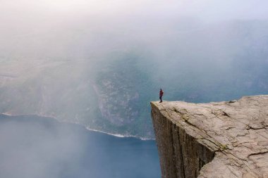 Norveç 'teki çarpıcı bir uçurumun kenarında tek başına duran bir yürüyüşçü, engin bir dağ manzarasına ve derin mavi sulara bakıyor..