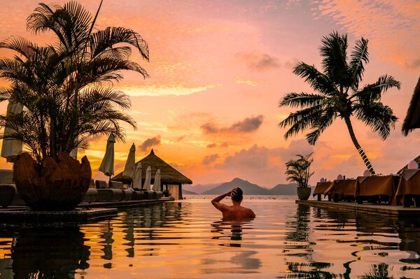 A young man enjoys a tranquil moment by a luxury swimming pool as the sun sets over the stunning Seychelles islands in La Digue. The serene environment invites relaxation and escape.