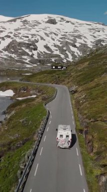 Norveç 'in göz kamaştırıcı güzelliğini tecrübe edin. Parlak mavi gökyüzünün altında yemyeşil ve görkemli kar örtülü dağlarla çevrili dolambaçlı bir yolda bir kampçı seyahat ederken..