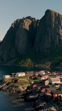 Büyüleyici köy, kıyı şeridi boyunca yüksek dağların altında geleneksel kırmızı kulübeler sergiliyor. Sakin sular doğanın güzelliğini yansıtır, Lofoten Norveç