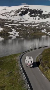 Norveç 'te karla kaplı görkemli dağlarla çevrili sakin bir göl kenarındaki dolambaçlı bir yolda bir karavan geziniyor. Sahne, Norveç güzelliğinin ve macerasının özünü yakalıyor..