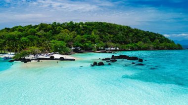 Canlı yeşil ağaçları ve yumuşak kumlu plajları olan yemyeşil bir adayı gösteren nefes kesici sahil manzarası. Canlı turkuaz sular, sakin bir tropikal atmosfer yaratır, Koh Kham Tayland