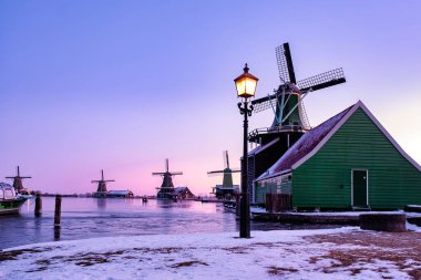 Zaanse Schans 'da kış alacakaranlığında ikonik yeşil ahşap evler ve geleneksel yel değirmenleri sergileniyor. Hollanda 'daki donmuş suyla zemini kaplayan kar, dingin atmosferi güçlendiriyor.